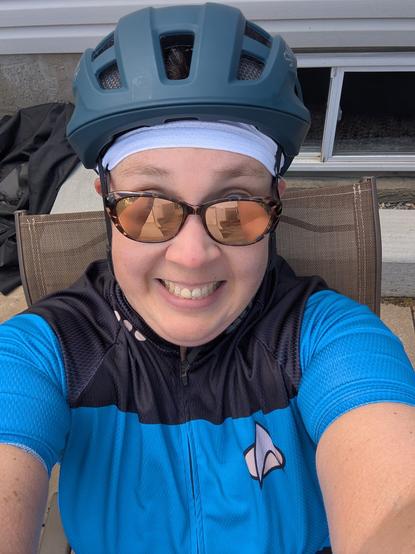 Exhausted and crazy looking cyclist sitting on a rocking lawn chair, wearing a teal helmet with a white sweatband peaking out underneath, brown tortoise shell sunglasses with gold reflective surface, cheesy grin, and a blue Star Trek The Next Generation jersey with 3 pips.