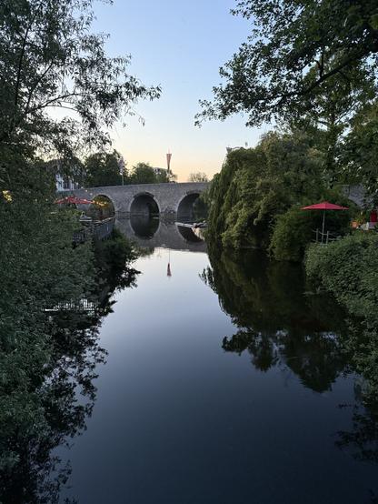 Das Bild zeigt eine ruhige Szene an einem Fluss, der von einem steinernen Bogenbrücken überspannt wird. Die Brücke hat mehrere Bögen, die sich in der glatten Wasseroberfläche spiegeln. Auf der linken Seite des Flusses sind Bäume und Büsche zu sehen, die den Flussrand säumen. Auf der rechten Seite befindet sich ein rotes Sonnenschirm, der einen Teil eines Outdoor-Raums markiert, möglicherweise eines Cafés oder Restaurants. Im Hintergrund ist ein Turm zu erkennen, der sich in den Himmel erhebt. Der Himmel ist klar und zeigt die sanften Farben des Abendhimmels. Die gesamte Szene ist von einer friedlichen und idyllischen Atmosphäre geprägt.
