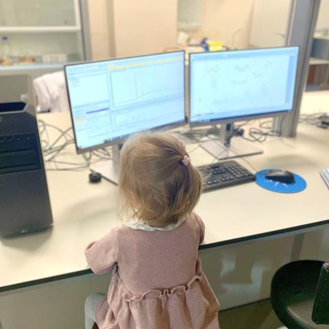 Ein kleines Kind sitzt auf einem Stuhl vor einem Laborarbeitsplatz und schaut aufmerksam auf den Bildschirm eines Computers, der mit einem Massenspektrometer verbunden ist.