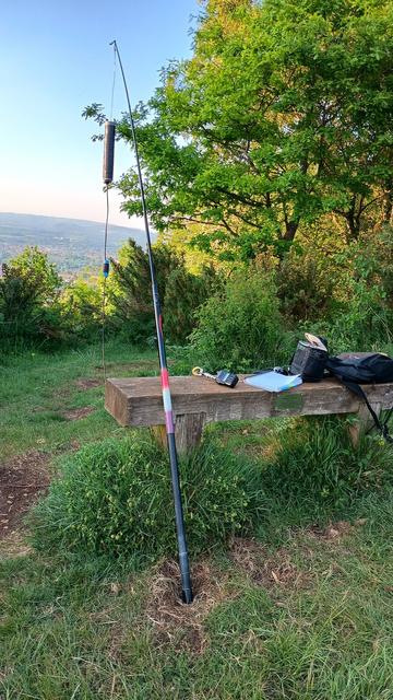 In the foreground is a handheld radio on a wooden bench, along with a notebook, case for the radio, and a backpack. Propped up beside the bench is a fishing rod from which hangs a flowerpot antenna. Behind the bench, and to the right of frame are some tall shrubs. In the distance is Cheltenham. The sky is blue.