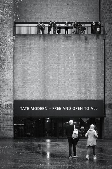Black and white. A man and a woman are walking towards the Tate Modern art gallery. It's a very tall brick-built former power station. There is a wide entrance with the words, "Tate Modern - Free and open to all" above it. High up, near the top of the picture is a balcony with a chest-height metal railing where eleven people are looking out at the view. One is currently looking at his phone, possibly researching St Paul's Cathedral which is almost opposite, across the River Thames.