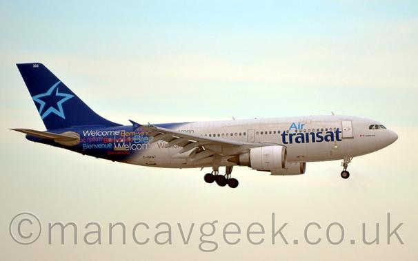Side view of a white and blue, twin engined jet airliner flying from left to right at a very low altitude, with undercarriage extended and flaps deployed from the rear of the wings, suggesting it is about to land.
The most of the body is white, with blue "Air Transat" titles on the forward fuselage.
The rear fuselage is a dark blue, with greetings in multiple languages, such as "Welcome", "Bienvenue", and "Wilkommen", printed in multiple colours.
The tail is also dark blue, with the blue outline of a star in the middle, a design repeated on the upturned wingtips.
Flat white sky fills the background.