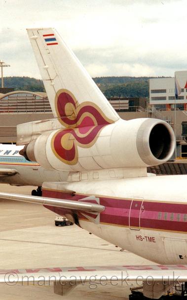 Close-up of the rear fuselage and tail of a 3 engined jet airliner parked facing to the right.
The plane is mostly white, with red and pink stripe outlined in gold running along the body, along with a red and pink flower design design also outlined in gold.
The ed registration "HS-TME" is on the lower rear fuselage.
A brown terminal building stretches across the frame in the background, with trees and rolling hills in the distance, under cloudy grey sky.