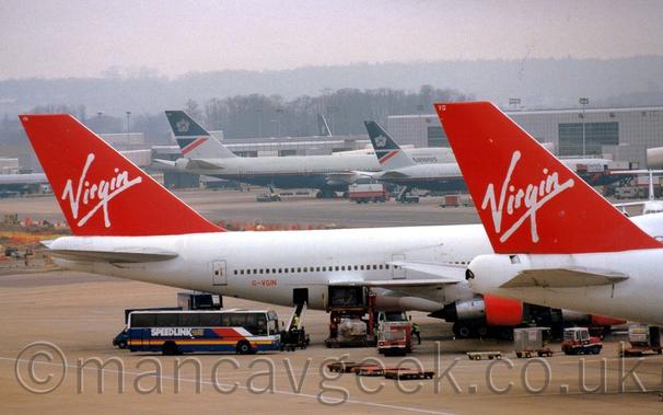 Close-up of the rear fuselage and tails of a pair of very large, white, 4 engined jet airliners parked facing to the right.
The planes are both mostly white, with red tails with white "Virgin" titles, and red engine pods.
The further plane has the red registration "G-VGIN" on the lower rear fuselage.
Baggage trucks, coaches, and other airport vehicles are scattered around the apron around them.
A grey  terminal building stretches across the background, with grey and dark blue airliners parked in front.
Trees slowly vanish into haze in the distance, under grim grey skies.