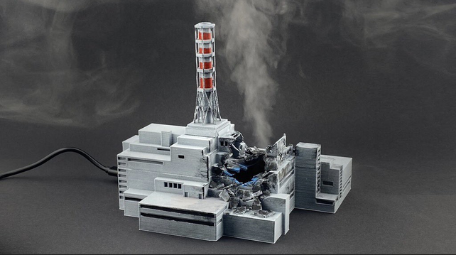 A miniature model of the damaged Chernobyl power plant, an industrial building with a tall chimney emitting smoke. The model is a detailed replica of the structure with visible destruction, smoke (humidity) rising from it and a black cable connected to it.