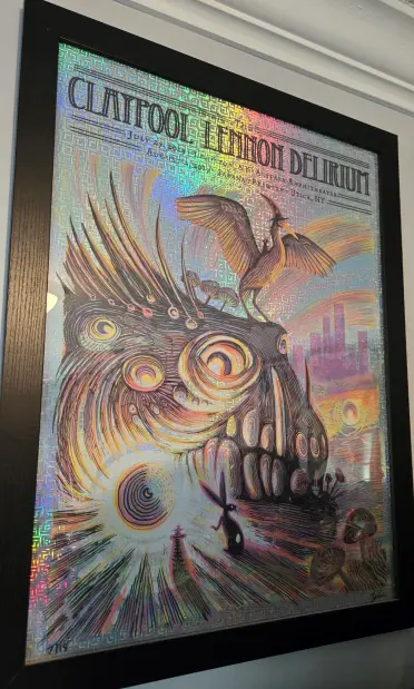 The image features a framed poster with a vibrant, psychedelic design. The title "CLAYFOOL LEMON DELIRIUM" is prominently displayed at the top in bold, black letters. Below the title, the dates "July 22, 2019 - August 1, 2019" and the location "Pocantico Amphitheater, Utica, NY" are written in smaller text. The background of the poster is a holographic pattern that reflects various colors, creating a shimmering effect.

The central illustration depicts a fantastical scene with a large, eye-like creature that has tentacle-like appendages. A bird with outstretched wings stands atop the creature, and a small black rabbit is positioned in the foreground, facing the creature. The background includes a cityscape with buildings and a sunset, adding depth to the scene. The overall design is intricate, with swirling patterns and a mix of organic and geometric shapes, contributing to the psychedelic aesthetic.