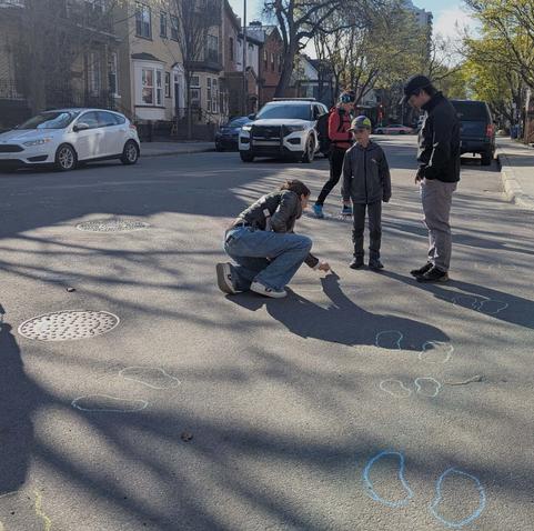 Dans une rue, une personne trace avec de la craie les contours des pas d'un jeunge garçon, accompagné d'un adulte. Une femme traverse la rue. En arrière-plan on aperçoit des voitures, dont ce qui semble une voiture de police. C'est une journée ensoleillée.
