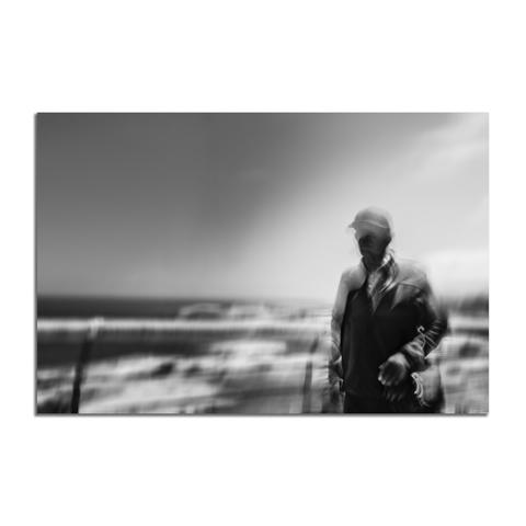 Mann an der Küste (ICM-Studie)

Die Schwarz-Weiß-Fotografie zeigt eine Küstenlandschaft mit einer Person im rechten Bilddrittel. Durch den Einsatz von Langzeitbelichtung und Intentional Camera Movement (ICM), einer bewussten Kamerabewegung während der Aufnahme, wirkt die Szenerie stark verwischt. Der Hintergrund, bestehend aus Himmel, Meer und Strand/Dünen, löst sich in horizontalen Streifen und weichen Flächen auf. Die menschliche Figur, erkennbar mit Kappe und Jacke, ist ebenfalls von Bewegungsunschärfe betroffen, hebt sich aber noch als dunkle Silhouette vom helleren Hintergrund ab. Die Technik erzeugt eine abstrakte, dynamische Darstellung der Szene, bei der Details zugunsten von Bewegung und Atmosphäre reduziert sind.