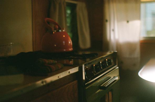 an orange kettle sits on top of an avocado green gas stove in a camper kitchen in diffused light.