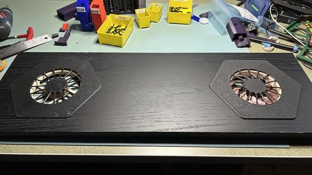 A black IKEA BILLY shelf has two large holes with hexagonal printed surrounds in them.  The right one holds a 120mm Noctua fan.  The left one holds a 3D printed thing that looks like a Noctua fan but with no blades.