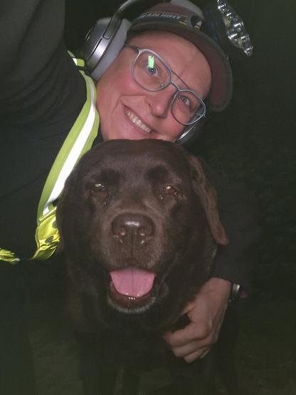 Night time selfie of a person and a chocolate brown Labrador with greying snout. The person is wearing a black jacket, yellow safety harness, red cap, silver headphones and blue-rimmed glasses. On their cap rests a headstrap torch in off position. Their free hand is around the dog's shoulders. Both are facing the camera. The dog's mouth is slightly open, showing off some blep. The person's head is resting on the dog's head. They're smiling, grateful for their time spent together.