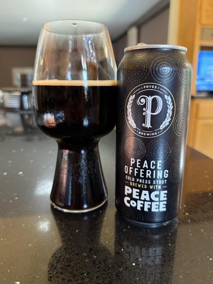A stout glass full of dark beer next to a black pint can with white lettering and a large scripted “P” in a circle in the middle