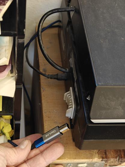 A hand holds a thin blue cable fitted with a much larger silver adapter in the cramped space behind a computer peripheral.