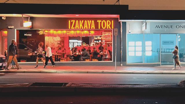 Pedestrians walk in front of a brightly lit restaurant that says, “Izakaya Tori” on a busy urban street