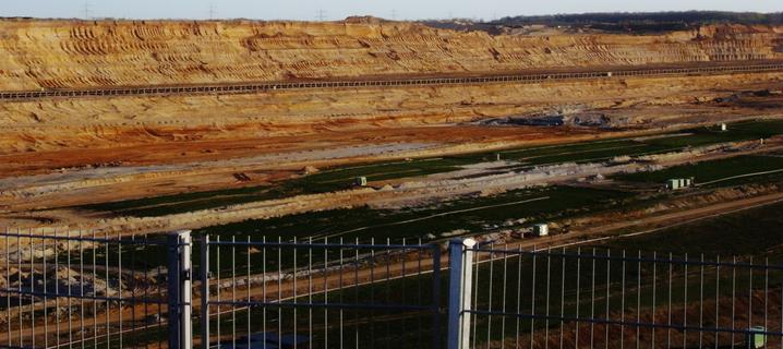 In the background, a small section of the Hambach open-cast mine's beige-orange-brown wall, dotted with uninterrupted horizontal lines, and a small part of the bottom of Germany's largest hole can be seen. It is also covered with lines (see alternative text of the middle image for more, please). On the left horizon, the tops of high-voltage pylons rise into the  sky, many treetops form the right horizon.
At the bottom of the image, more leading lines can be seen in the form of many thin horizontal and some vertical bars of a metal fence. There is also some turf.