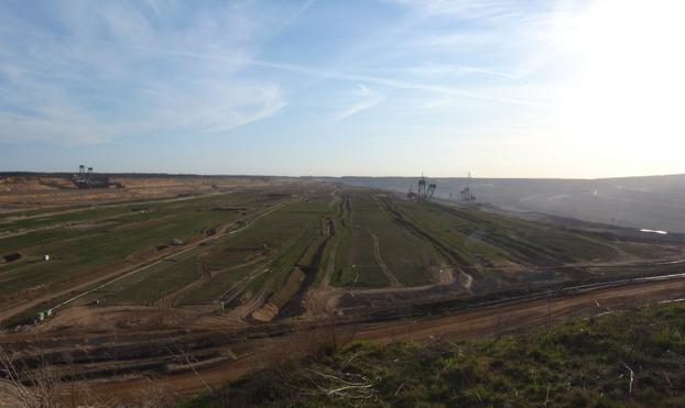 Below the right horizon, more of the wall from the left image can be seen; most of the image is taken up by a section of Germany's largest hole. Conveyor belts and channels for the gigantic excavators, which look so tiny in the image because the hole surrounding them is so vast. To the right, an even deeper section stretches out, covered in haze.
So far away that they are barely visible, treetops and the tops of power pylons form the horizon.
A blue sky stretches over the hole, covered by white, partly melting scars left by airplanes. At the bottom of the image, there is some turf.