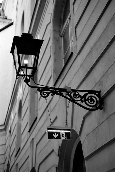 The black and white image shows an ornate metal street lamp mounted on the side of a building. Hanging beneath it is a modern illuminated emergency exit sign with a figure running toward a downward arrow, creating a contrast between old and new elements.