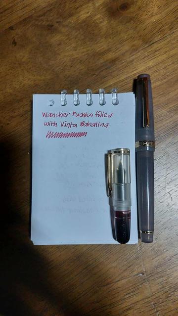 A tiny transparent pen is on an A7 notepad with a writing sample, next to a bigger pen. It is half the size of the pen.