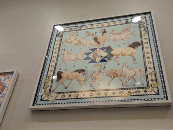 Blue scarf framed on the wall of a bunch of horses running around on light blue. Bays, Palominos and Grey horses with a yellowish border and blue border