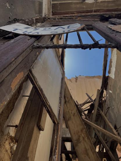 Interior of a ruined house. Looking up through a broken front door with half a For Sale sign on it, torn, phone number illegible, past the fallen floor boards and the roofless walls, to the clear blue sky.