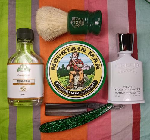 (Le Chat Mistral description)

The image showcases a collection of grooming items arranged on a striped background. At the centre, there's a round tin of shaving soap labelled "Mountain Man" from Stirling Soap Company. To the left, there's a small bottle of aftershave splash, also labelled "Mountain Man." On the right, there's a bottle of "Silver Mountain Water" by Creed. Above the soap tin, there's a shaving brush with a green handle and a white bristle. Below the soap tin, there's a straight razor with a green, glittery handle. The items are neatly laid out, suggesting a traditional shaving setup.