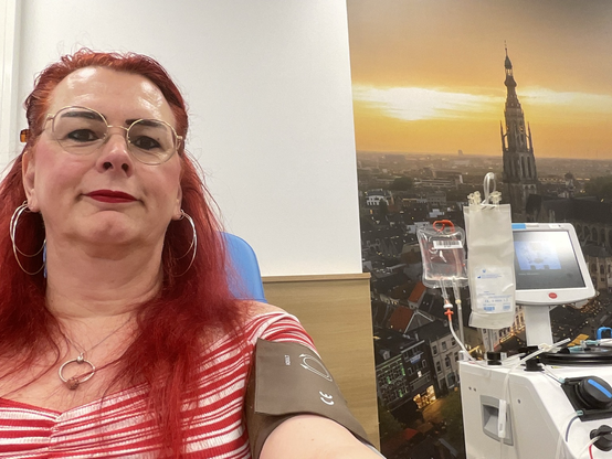 Emma, met rood haar en bril, glimlacht ontspannen tijdens haar plasmadonatie bij de bloedbank. Achter haar zijn medische apparaten te zien die gebruikt worden voor het afnemen van plasma. Ze draagt een wit met rood gestreepte off-shoulder top.