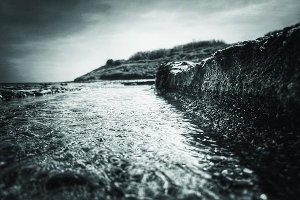 A low-angle, monochrome photograph taken at ground level on a beach shows shallow water flowing over a small, carved-out sandbank towards the sea under a cloudy sky.