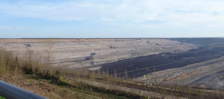 Several excavators, really giant ones that look tiny in the hole, are doing their work.In the foreground, a small white-flowering fruit tree and several leafless bushes can be seen, as well as a small part of the upper edge of a fence, a green-painted steel pipe. A white-blue sky arches above the pit.