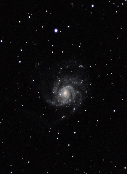 Fotografie eines sternklaren Nachthimmels mit der Spiralgalaxie Messier 101 im Zentrum. Die Galaxie zeigt eine helle Mitte und locker gewundene Spiralarme, durchsetzt mit hellen Sternentstehungsgebieten. Umgeben ist sie von zahlreichen Einzelsternen vor schwarzem Hintergrund.