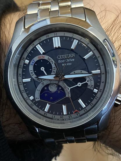 Un reloj de pulsera analógico marcando las 9:15. El reloj tiene tres complicaciones pequeñas que marcan el mes, el día de la semana y la fase actual de la luna. El día del mes se marca con una aguja más larga que va señalando números del 1 al 31 situados en la parte más exterior del dial.