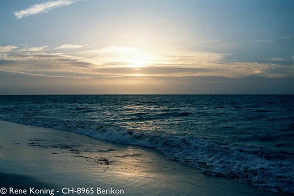 The image depicts a serene beach scene at sunset. The sky is painted with soft hues of blue and orange, with the sun partially visible near the horizon, casting a warm glow over the scene. Wispy clouds are scattered across the sky, reflecting the sunlight. The ocean is calm, with gentle waves lapping at the shore, creating a foamy white edge where the water meets the sand. The beach is mostly empty, with a few scattered rocks and debris visible on the wet sand. The overall atmosphere is tranquil and peaceful, with the natural beauty of the sunset and the ocean dominating the scene.