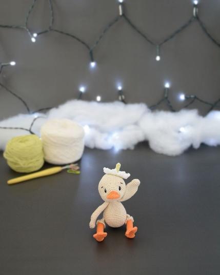Amigurumi représentant un caneton crème qui fait coucou avec une aile. Il a un bec et des pattes orange, une fleur blanche sur la tête. Il est assis devant deux pelotes de coton, un crochet et une guirlande lumineuse en arrière-plan.