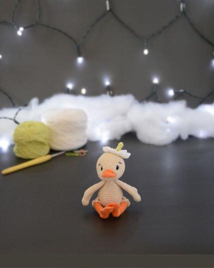 Amigurumi représentant un caneton crème assis. Il a un bec et des pattes orange, une fleur blanche sur la tête. Il est devant deux pelotes de coton, un crochet et une guirlande lumineuse en arrière-plan.