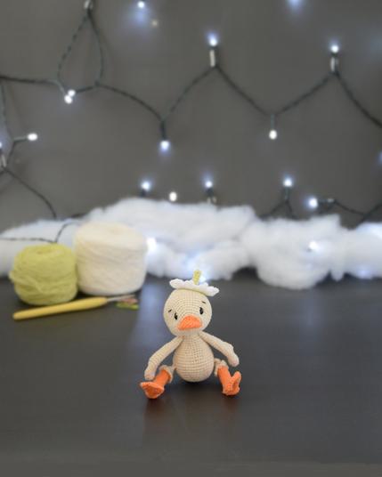 Amigurumi représentant un caneton crème assis. Il a un bec et des pattes orange, une fleur blanche sur la tête. Il est devant deux pelotes de coton, un crochet et une guirlande lumineuse en arrière-plan.