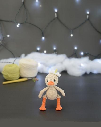Amigurumi représentant un caneton crème debout. Il a un bec et des pattes orange, une fleur blanche sur la tête. Il est devant deux pelotes de coton, un crochet et une guirlande lumineuse en arrière-plan.