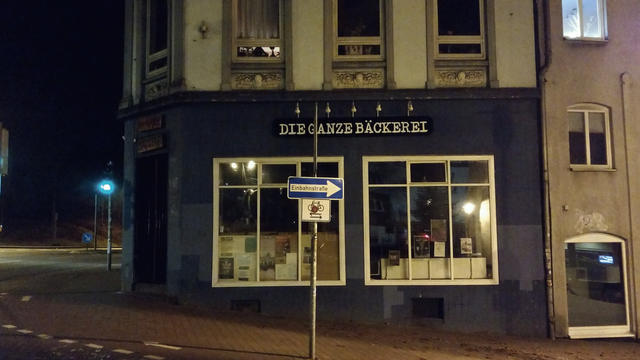 Night shot of a dark facade of a residential building. The lower floor houses a bakery, recognizable by the writing above the windows saying DIE GANZE BÄCKEREI in German, translates as ALL OF THE BAKERY. Overall photo s a dark-ish night shot of the building at a street corner, the black exterior walls and early 1900 facade decor.