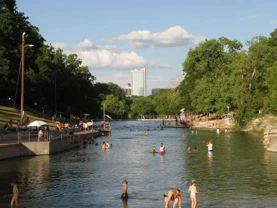 The image depicts a scenic river or waterway lined with trees. There are several people swimming and enjoying the water, with some wading in the shallows. In the background, you can see tall buildings, suggesting an urban setting nearby. The sky is partly cloudy, creating a pleasant atmosphere for outdoor activities.

Image Credits: Wikimedia / Downtown Austin / Public domain