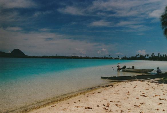 Plage de sable fin et mer bleu azur dans le lagon, avec des pirogues sur la droite et des montagnes volcaniques au loin sur la gauche, sur l'île de Huahine en Polynésie française.