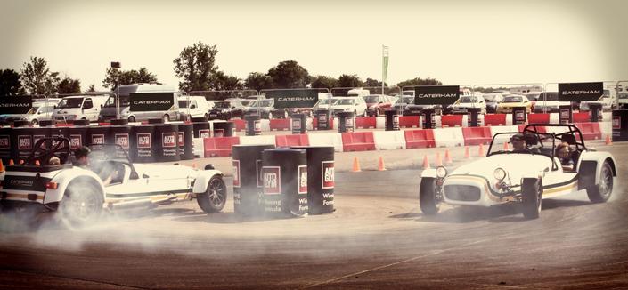 Two Caterham sports cars drifting in a closed track area, producing tyre smoke. Both cars are white with black and yellow detailing. Drivers are accompanied by passengers wearing helmets. The track is marked with orange cones and black barrels.