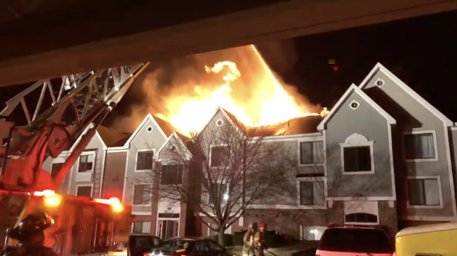 Apartment building engulfed in flames at night. Firefighters and a firetruck with a ladder are on the scene. The atmosphere is urgent and intense.