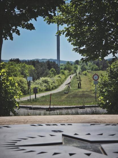 The image displays a path traversing a grassy area, bordered by trees and bushes. Numerous dark, abstract metal sculptures are positioned along the path. The path surface is light-colored. To the right of the path, a round sign with a red border signaling that vehicle traffic along the path is not allowed. In the distance, a tall, slender tower with a platform is visible against a clear sky, with rolling, tree-covered hills in the background. A signpost with multiple signs stands to the left of the path. Several figures are walking on the path in the distance. The foreground contains a textured surface with star-shaped cutouts-a compass rose. Based on the visible portion of the compass rose in the foreground, the camera's perspective is suggested to be facing southwest.