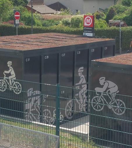 Das Bild zeigt eine Reihe von schwarzen, rechteckigen Parkboxen, die als Fahrradparkplätze dienen. Jede Box ist mit einer weißen Zahl gekennzeichnet, wie z.B. C 31, C 29, C 27, C 25, C 23 und C 21. Auf den Boxen sind weiße Silhouetten von Radfahrern abgebildet, die verschiedene Fahrradarten und Positionen darstellen. Die Boxen sind mit einer grünen Metallzaun umgeben, der einen Bereich abgrenzt. Im Hintergrund ist ein Schild mit einem roten "M" und der Aufschrift "Mobilisation" zu sehen, was auf eine Nahverkehrsstation hinweist. Ein weiteres Schild zeigt die Straße "Finkenberger Straße". Die Umgebung ist grün und gepflegt, mit Bäumen und Sträuchern.