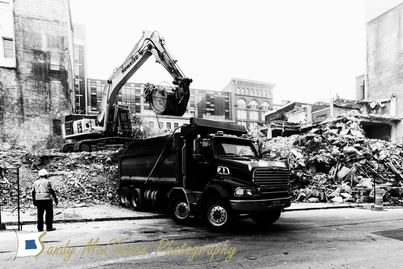 Excavator sitting on a pile of demolition debris loads a dump truck in the downtown of a city.