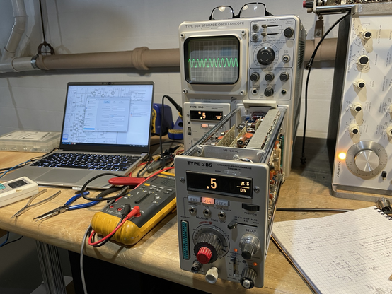 A workbench with a laptop, multimeter, hand written notes, and an oscilloscope with its horizontal plug-in in the service position on an extender. A clean sine wave is displayed on the screen from a signal generator on the right.