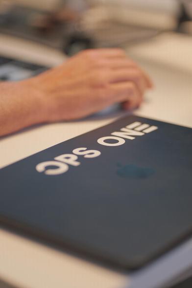 Close-Up von einem Bürotisch, im Hintergrund eine Hand, die eine Computermaus bedient, im Vordergrund ein Laptop mit Ops-One-Kleber.
