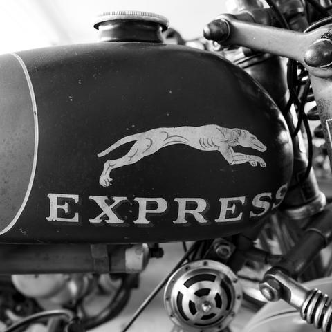 Eine Teilaufnahme von einem alten Motorrad. Auf dem Tank sieht man einen weißen Windhund auf dunkel grauem Hintergrund. Darunter steht in Großbuchstaben EXPRESS. Man sieht außerdem die Hupe und einen kleinen Teil der Gabe. Das Foto ist in Schwarzweiß.