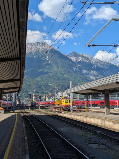 Scenery of Innsbruck Hbf station: A parked yellow catenary maintenance vehicle in front of an eastbound Railjet 2 long distance train. On the left another westbound Railjet arriving at the platform. In the backgrounf buildings of the city, including a church tower, and tall green and rocky Alps mountains