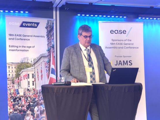 Cem Uzun at the podium of the EASE AGM. He's a Turkish man with short greying hair, wearing glasses and a grey suit and checked tie.