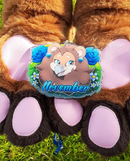 A close-up photograph of a large acrylic charm badge with artwork of a brown female gryphon surrounded by a bush of blue roses and smaller blue flowers. The charm is being held in the paws of a fursuit resembling the character in the badge. The charm measures 6 in. (15.24 cm) on its longest side, making it very visible from a distance.