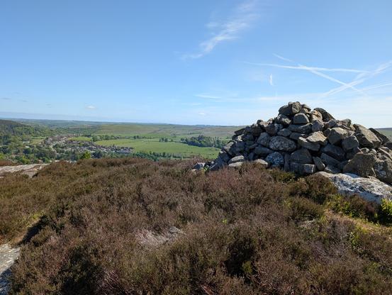 A large cairn is in the right of the image. To the left are views of green fields and the small town of Rothbury.